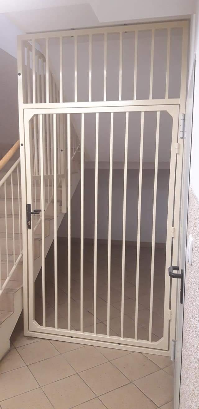 Beżowa metalowa brama z pionowymi prętami zabezpieczająca wejście na klatkę schodową wewnątrz budynku.