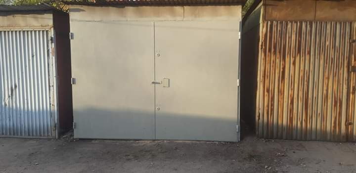 Szare dwuskrzydłowe metalowe drzwi garażowe pomiędzy dwoma starszymi garażami z blachy falistej, widoczne ślady korozji.