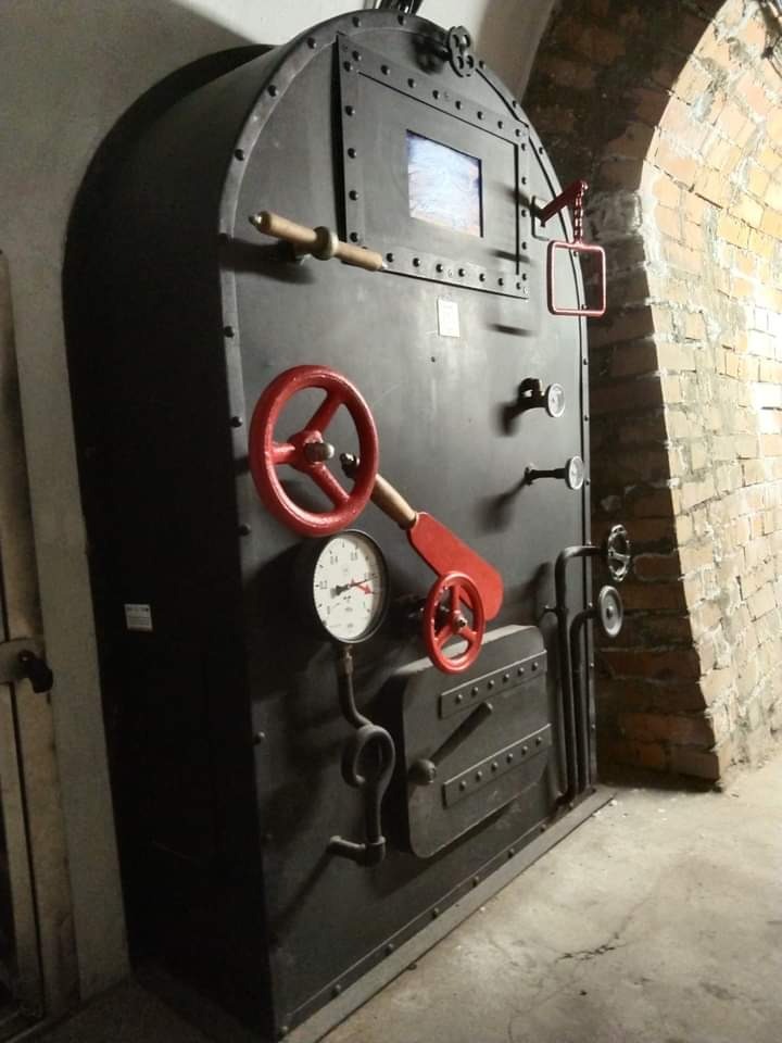 Ciężkie, metalowe drzwi z okienkiem i czerwonymi elementami sterującymi, umieszczone w ceglanym łuku.