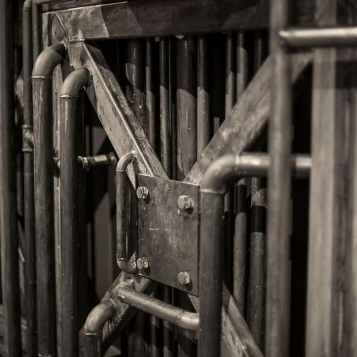 Fragment metalowej konstrukcji bramy z elementami rurowymi, z widocznymi spawami i śrubami, w odcieniach szarości.