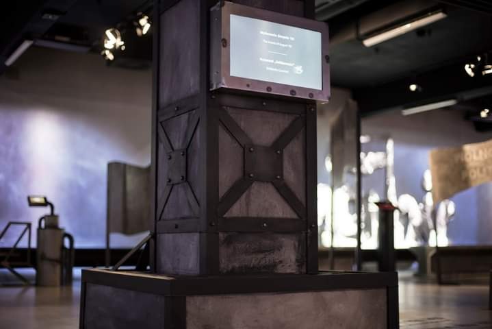 Metalowa konstrukcja przypominająca filar z dekoracyjnymi elementami krzyżowymi i ekranem wyświetlającym tekst w muzeum lub galerii.