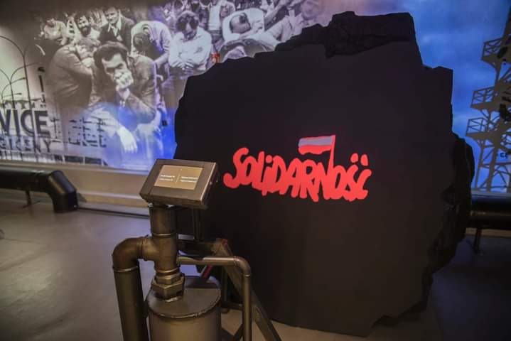 Instalacja muzealna: fragment ściany z napisem 'Solidarność' w czerwonej czcionce i flagą Polski, obok metalowa rura z zaworem i ekran dotykowy, w tle zdjęcia z epoki i konstrukcja wieżowa.