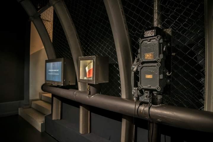 Metalowa instalacja z rurami, skrzynkami i wyświetlaczami na tle siatki, w industrialnym wnętrzu.