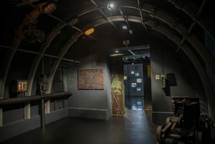 Wnętrze stylizowanej piwnicy w Rudzie Śląskiej z metalową konstrukcją łukową, rurami na ścianach i obrazem w tle, imitujące podziemny schron.