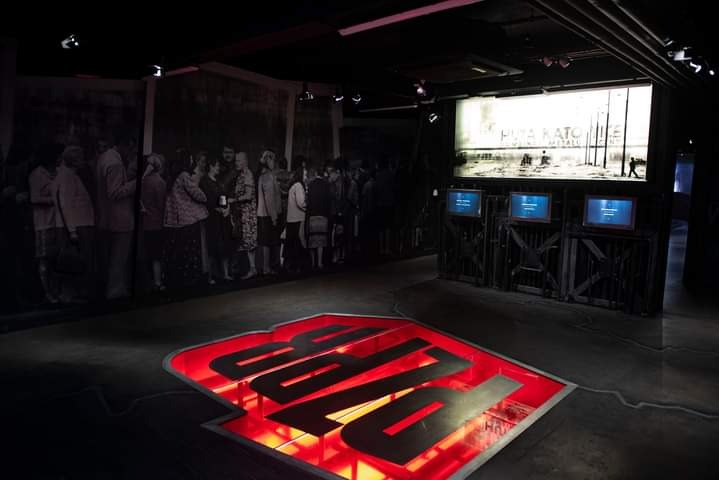 Wnętrze muzeum z podświetlanym logo na podłodze, czarno-białym muralem postaci w tle i trzema monitorami prezentującymi informacje.