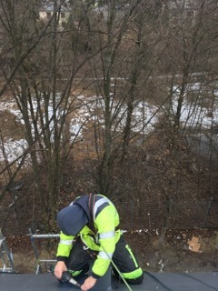 Pracownik firmy z Katowic w jaskrawej odzieży roboczej, w trakcie pracy na dachu, używa wkrętarki. W tle widoczne drzewa i zabudowania.