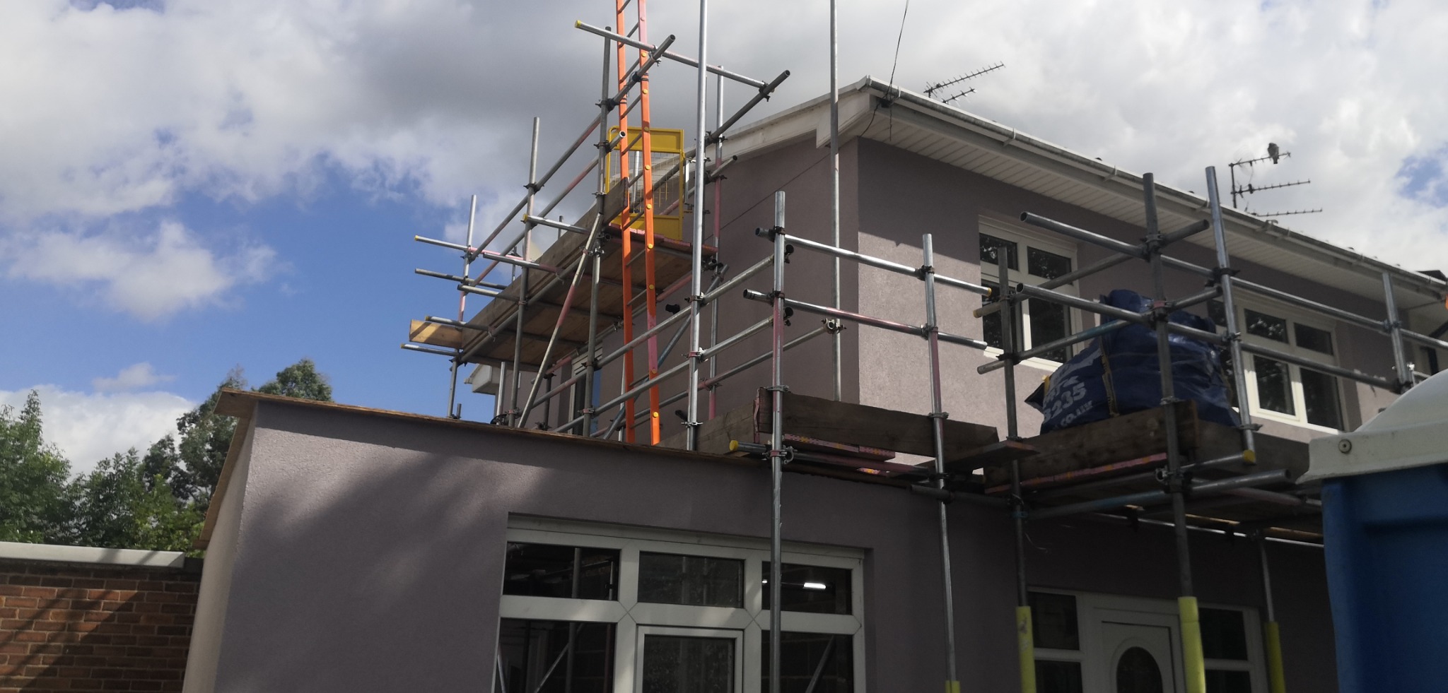 Dom jednorodzinny w trakcie prac elewacyjnych, widoczne rusztowania, świeżo położony tynk w odcieniach szarości, niebieski worek z materiałami budowlanymi na rusztowaniu.