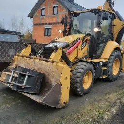 Żółta koparko-ładowarka Caterpillar 432F2 z łyżką załadowaną czarnym kontenerem na tle ceglanego budynku w trakcie budowy.