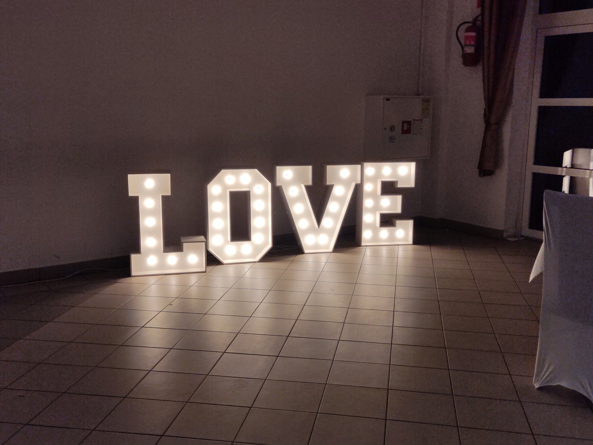 Podświetlany napis LOVE z żarówkami na podłodze z płytek, widoczny fragment krzesła z białym pokrowcem, w tle ściana z gaśnicą i zasłonięte okno.