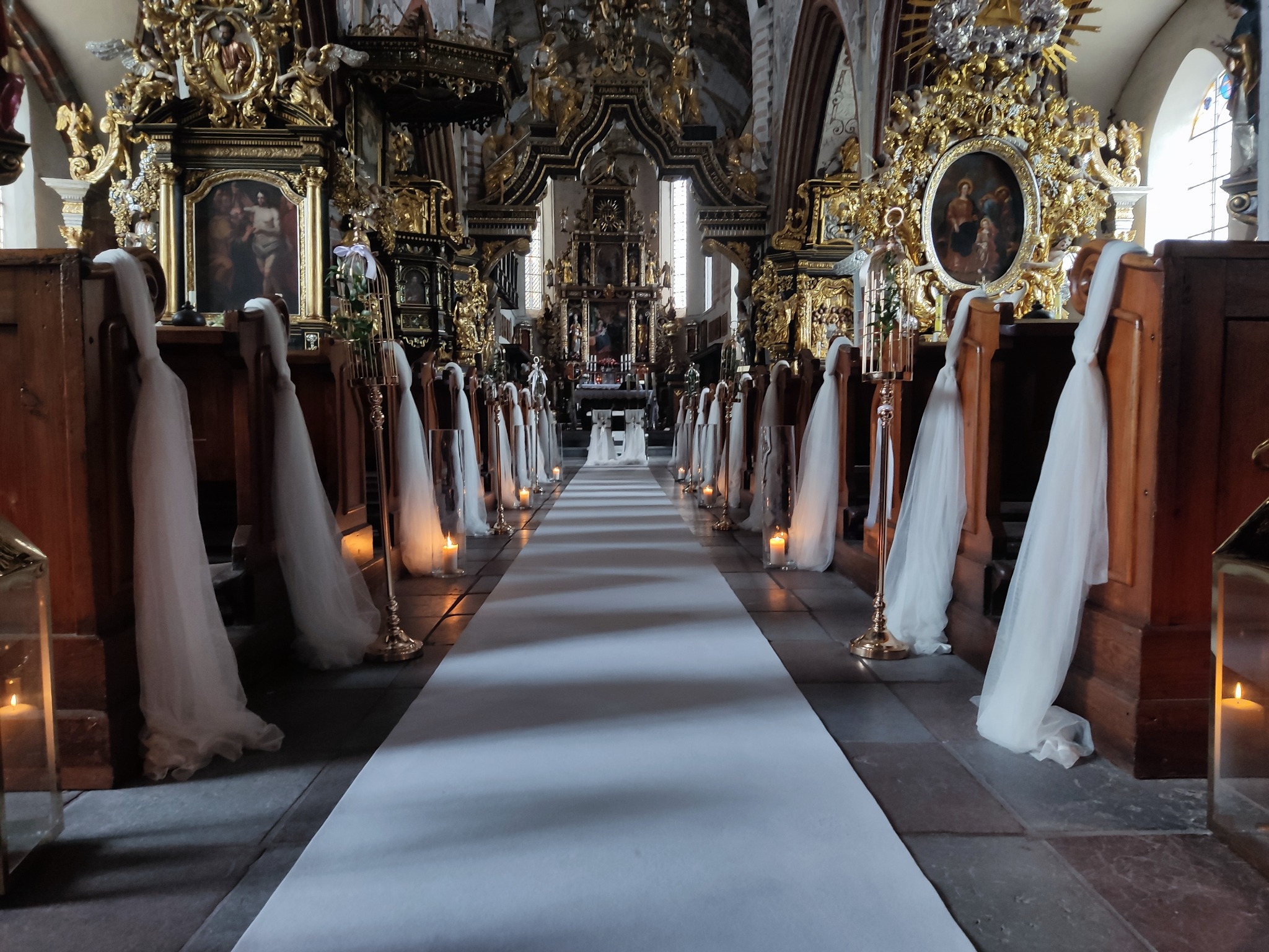 Uroczystość ślubna w bogato zdobionym kościele: biały dywan prowadzący do ołtarza, ławki udekorowane białymi tiulowymi wstążkami, świece w złotych lampionach, złote zdobienia ołtarza i ścian.