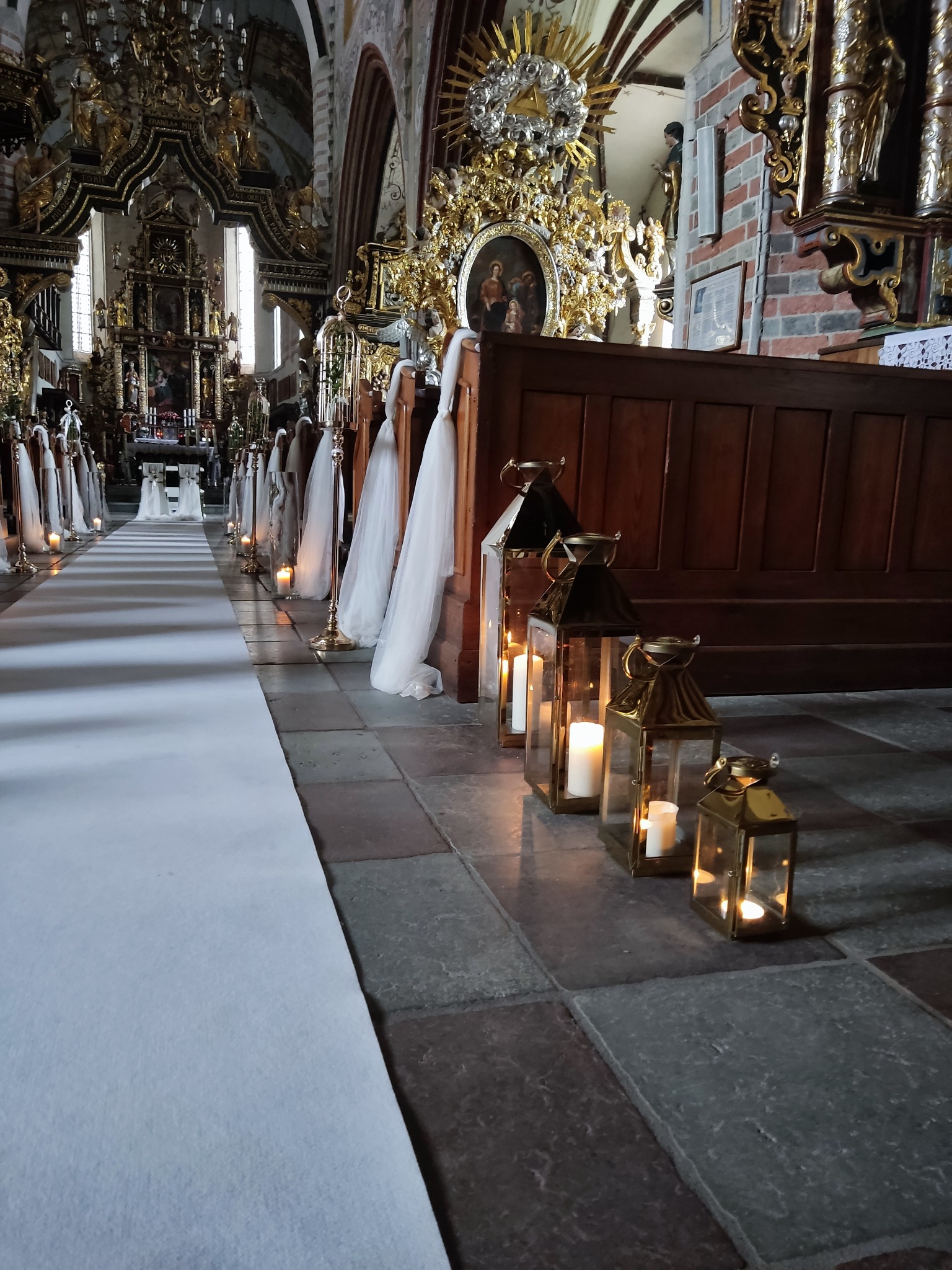 Udekorowane przejście w kościele na uroczystość: biały dywan, lampiony ze świecami i tiulowe wstążki na ławkach.