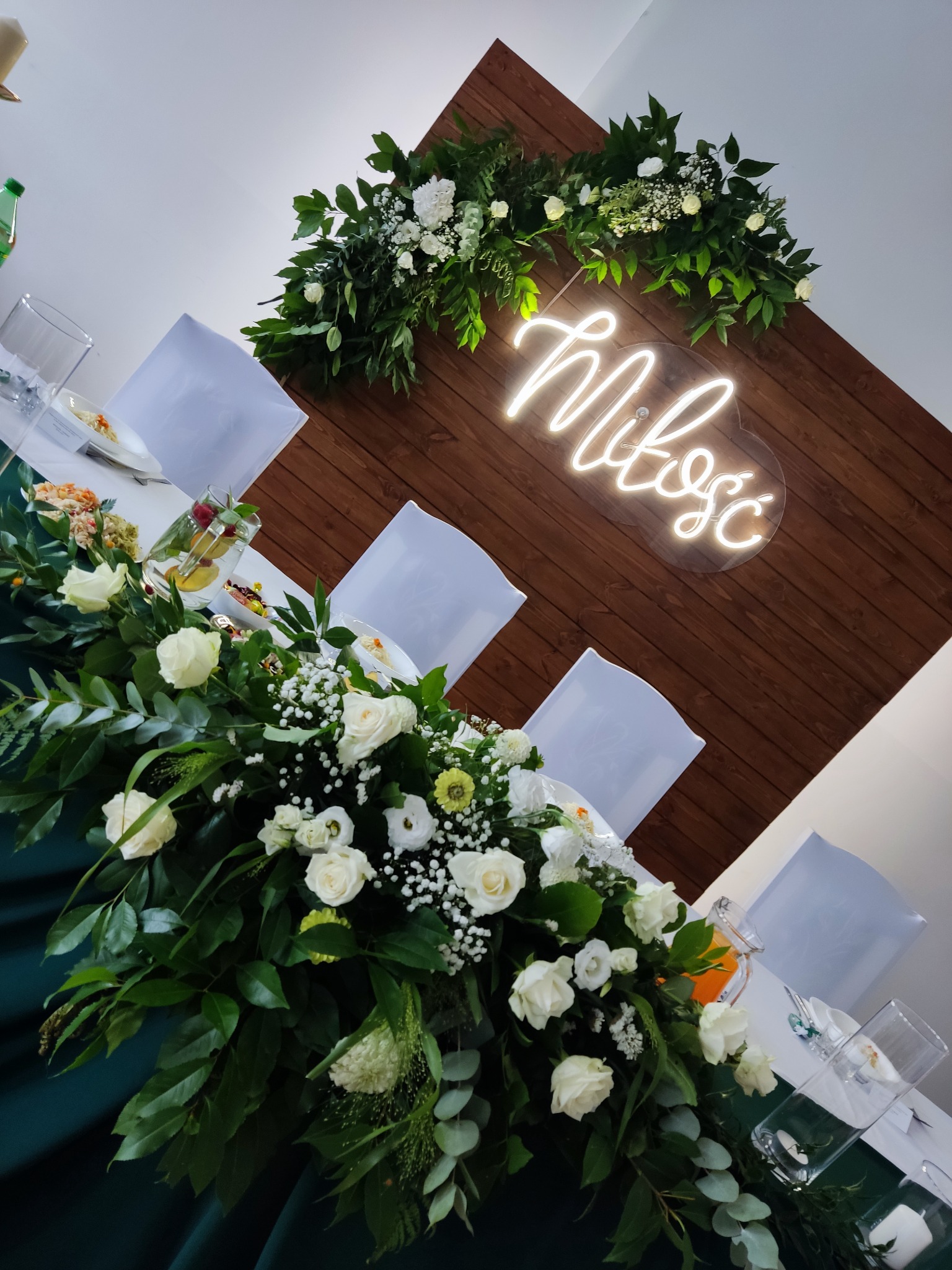 Udekorowany stół weselny z białymi różami, zielonymi liśćmi i neonowym napisem 'Miłość' na drewnianym tle.