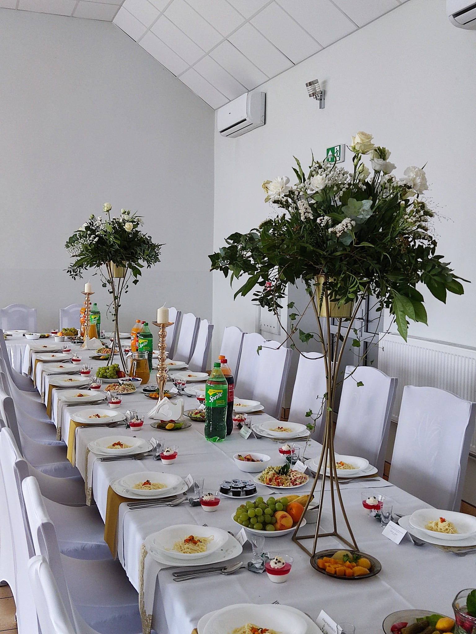 Uroczysty stół weselny z białymi pokrowcami na krzesłach, złotymi świecznikami i wysokimi kompozycjami kwiatowymi w złotych wazonach, zastawiony talerzami z makaronem, owocami i napojami.