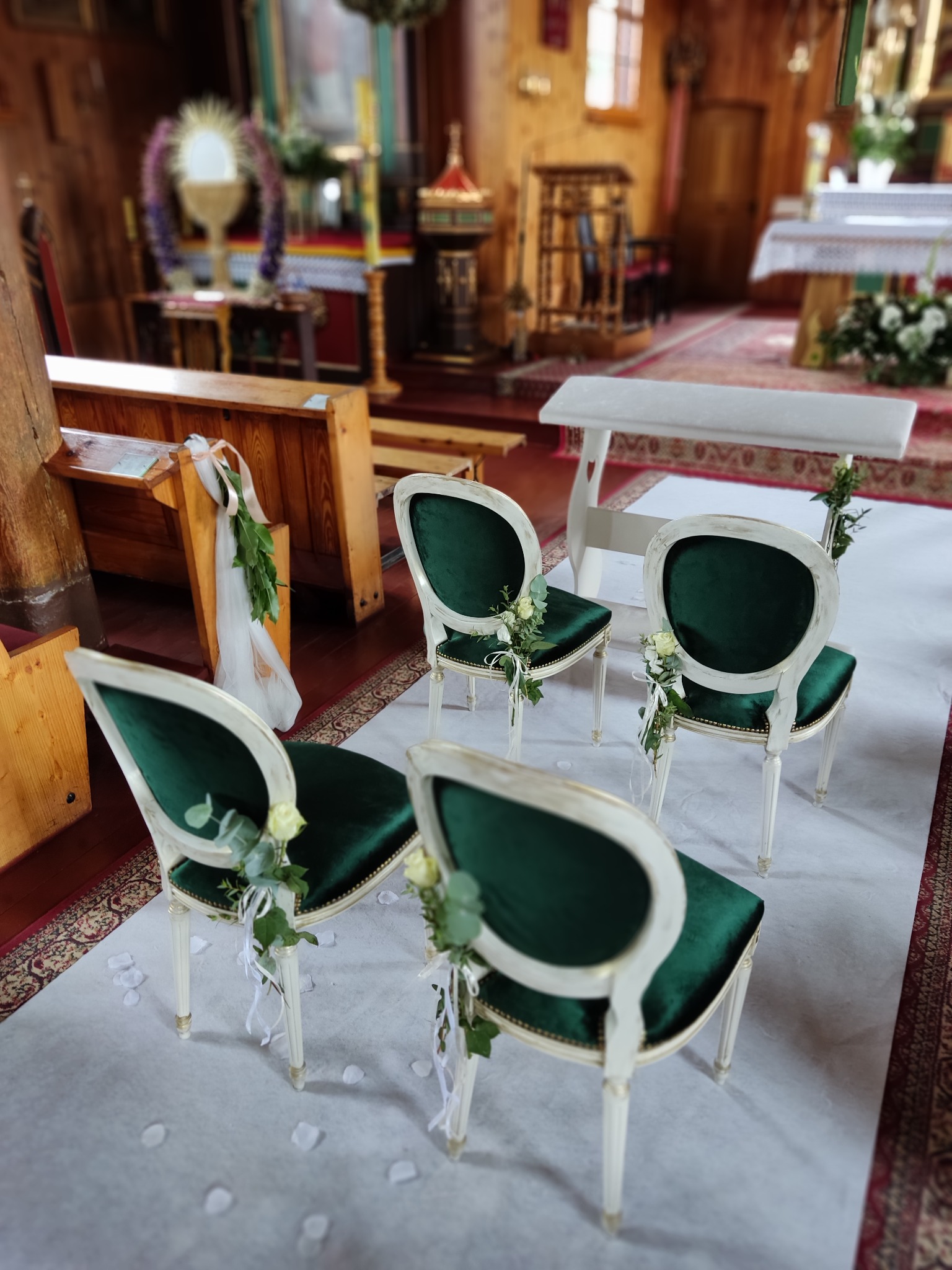 Uroczystość w kościele: Cztery krzesła z zielonym aksamitem i białymi ramami, udekorowane białymi różami i wstążkami, ustawione na białym dywanie wzdłuż nawy głównej.