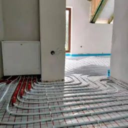 Montaż ogrzewania podłogowego w surowym wnętrzu: białe rury ułożone na czerwonej siatce, widoczne elementy instalacji i fragment okna.