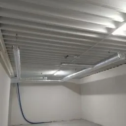 Surowy stan pomieszczenia z widocznymi kanałami wentylacyjnymi pod sufitem i niebieskim przewodem przy ścianie.