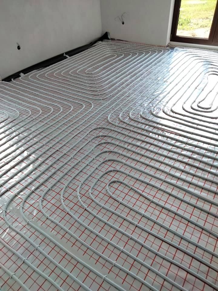 Białe rurki ułożone w pętle na podłodze z czerwono-białą kratką, tworzące system ogrzewania podłogowego, widok z góry, surowy stan wykończenia wnętrza.