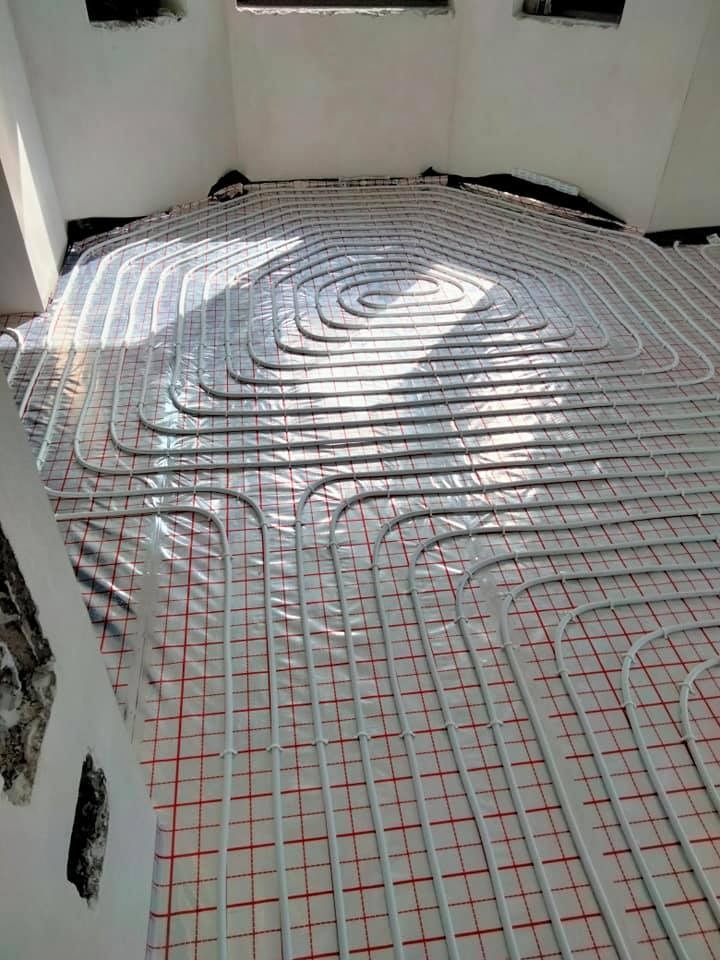 Białe rury ułożone spiralnie na folii z czerwoną kratką, instalacja przed wylewką, widoczne fragmenty surowych ścian w pomieszczeniu.