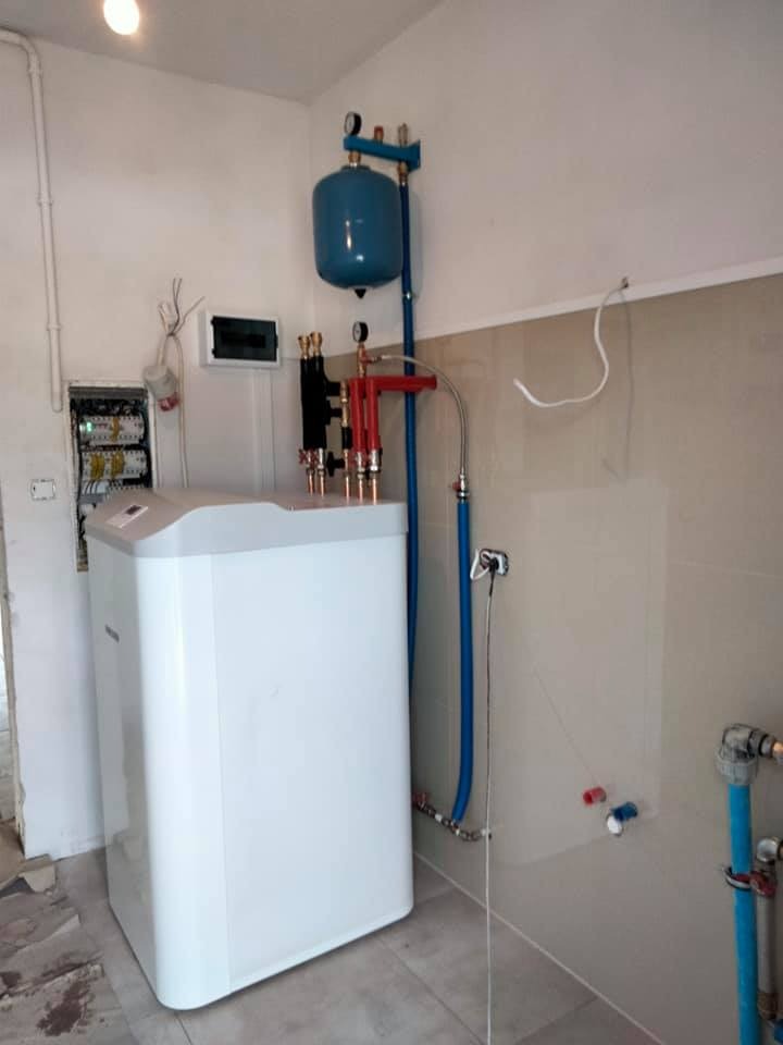 Biała jednostka centralna z niebieskim zbiornikiem wyrównawczym, czerwonymi i czarnymi zaworami, oraz niebieskimi rurami w pomieszczeniu z płytkami ściennymi, instalacja w trakcie podłączania.