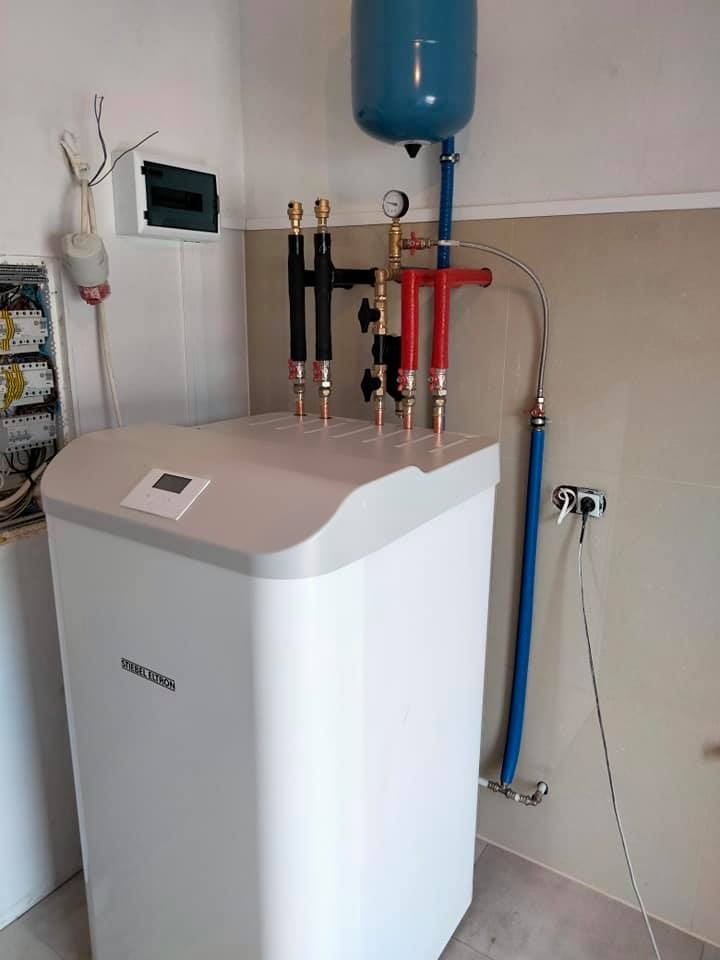 Biała jednostka grzewcza Siebel Eltron z niebieskim zbiornikiem wyrównawczym, instalacją hydrauliczną w kolorach czarnym i czerwonym, zamontowana w pomieszczeniu z beżowymi płytkami.