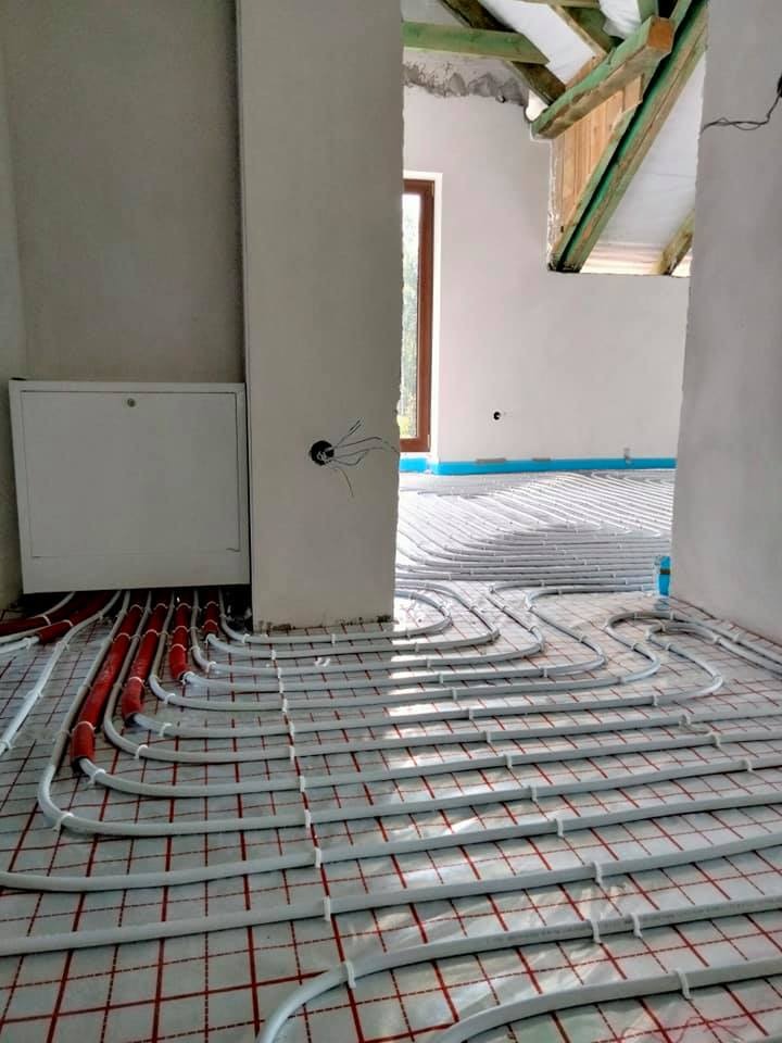 Montaż ogrzewania podłogowego w surowym wnętrzu: białe rury ułożone na czerwonej siatce, widoczne elementy instalacji i fragment okna.