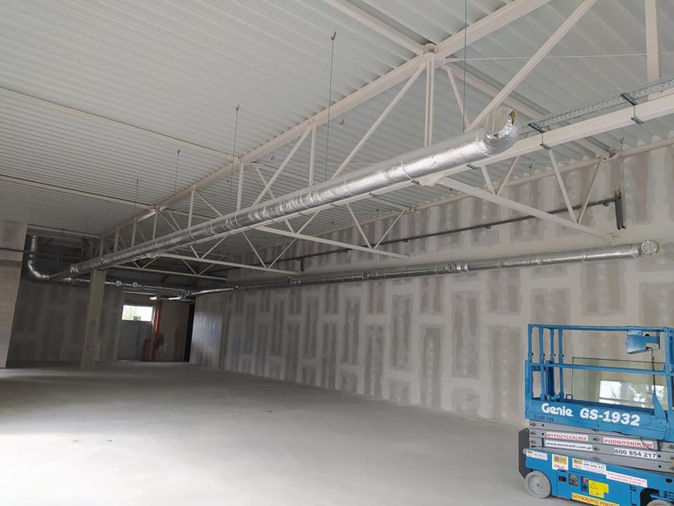 Wnętrze hali w trakcie montażu instalacji wentylacyjnej z widocznymi srebrnymi rurami pod sufitem i podnośnikiem Genie GS-1932 w prawym dolnym rogu.