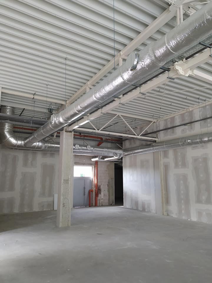 Wnętrze komercyjnego budynku w trakcie instalacji systemu wentylacji z widocznymi, zaizolowanymi przewodami wentylacyjnymi pod sufitem i ścianami przygotowanymi do wykończenia.