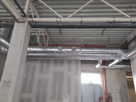 Instalacja przewodów wentylacyjnych w budynku komercyjnym, widoczne rury w izolacji, surowa ściana z płyt gipsowych i stalowa konstrukcja dachu.