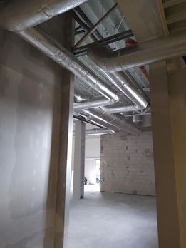 Widok wnętrza w trakcie instalacji systemu wentylacyjnego z widocznymi srebrnymi rurami pod sufitem, w budynku w Pilawie. Surowe ściany z betonu komórkowego i kolumny konstrukcyjne.