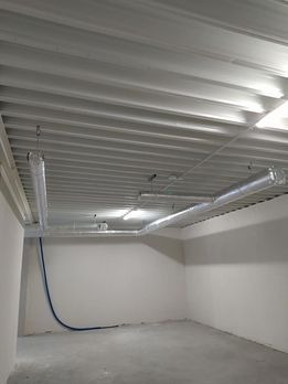 Surowy stan pomieszczenia z widocznymi kanałami wentylacyjnymi pod sufitem i niebieskim przewodem przy ścianie.