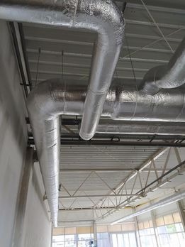 Izolowane kanały wentylacyjne z widoczną aluminiową folią, zawieszone pod sufitem hali przemysłowej, widok z dołu.