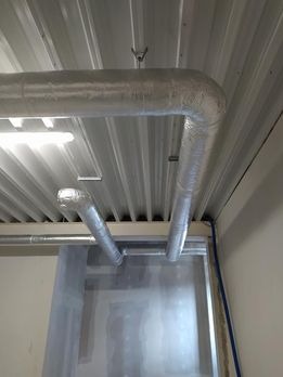 Metalowe rury wentylacyjne w izolacji termicznej, zamontowane pod sufitem z blachy trapezowej w pomieszczeniu przemysłowym, widoczne elementy mocujące.
