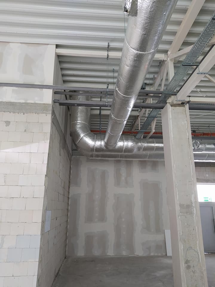 Instalacja wentylacyjna z izolacją aluminiową, widoczne rury pod sufitem w surowym wnętrzu budynku komercyjnego z nieotynkowanymi ścianami i betonową podłogą.