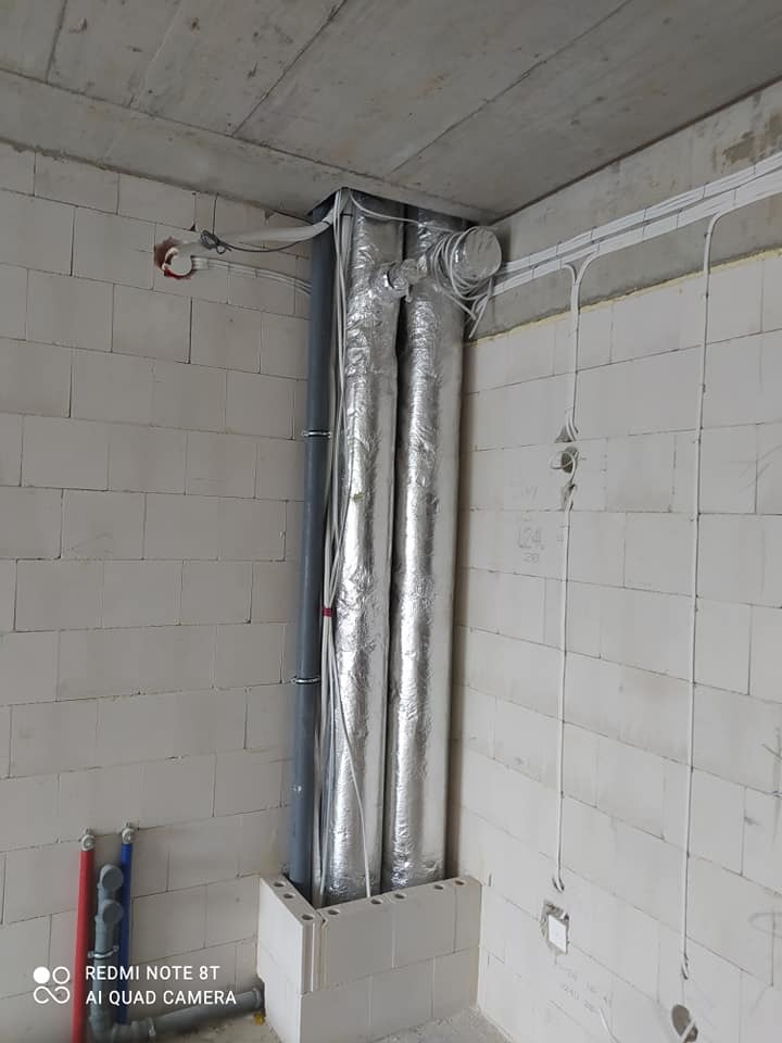 Dwa pionowe przewody wentylacyjne w srebrnej izolacji, prowadzone w narożniku pomieszczenia z białych bloczków, widoczne także rury instalacji wodnej i kable elektryczne.