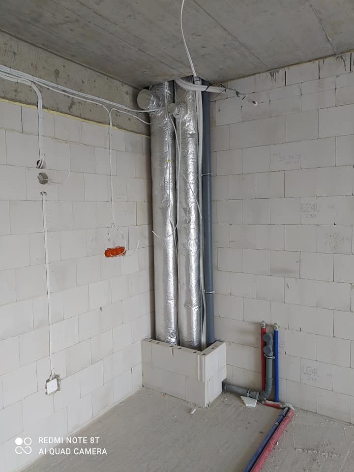 Surowy stan pomieszczenia z widocznymi pionami instalacji wentylacyjnej w srebrnej folii izolacyjnej, rurami kanalizacyjnymi i instalacji wodnej. Ściany z białego betonu komórkowego, częściowo...