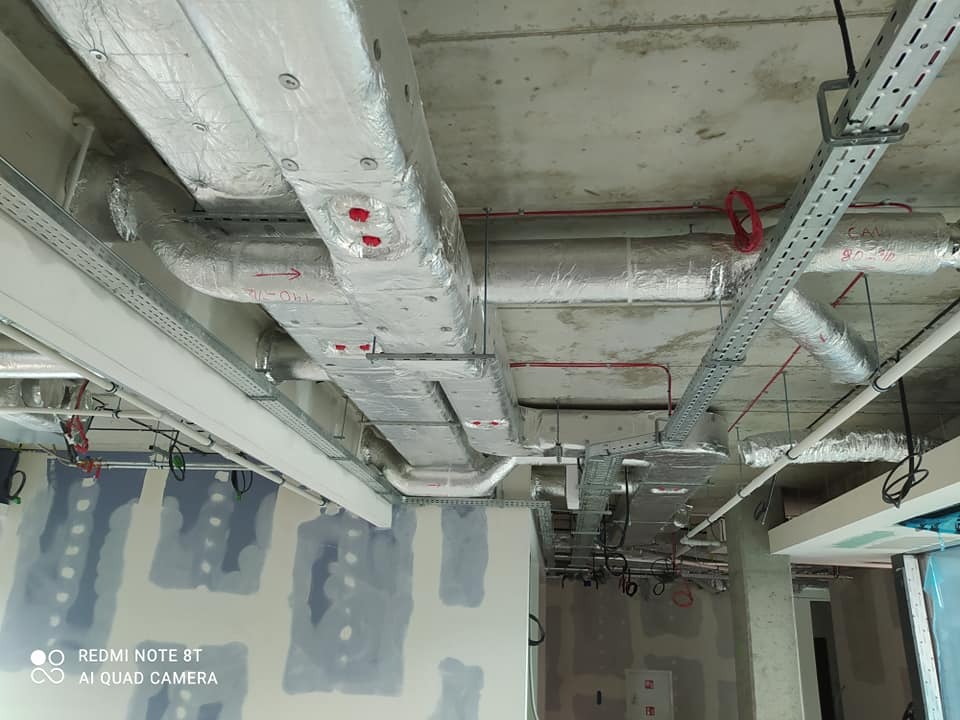 Instalacja wentylacyjna z izolowanymi kanałami na suficie w surowym stanie, widoczne koryta kablowe i niedokończone ściany z płyt gipsowo-kartonowych.