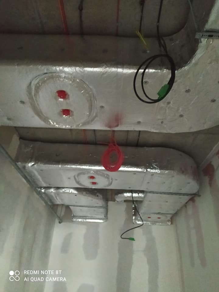 Widok od dołu na izolowane kanały wentylacyjne w surowym wnętrzu, widoczne przewody elektryczne i fragment ściany w trakcie wykończenia.