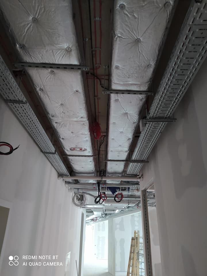 Montaż instalacji wentylacyjnej i elektrycznej pod sufitem w budynku komercyjnym, widoczne ocieplone kanały wentylacyjne, koryta kablowe i przewody elektryczne.