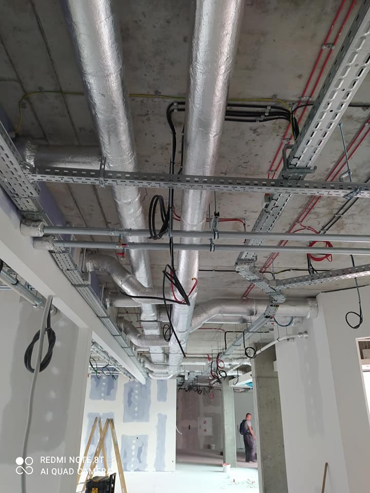 Instalacja przewodów wentylacyjnych z izolacją aluminiową i tras kablowych pod sufitem w surowym stanie budynku, widoczne drabiny i pracownik w tle.