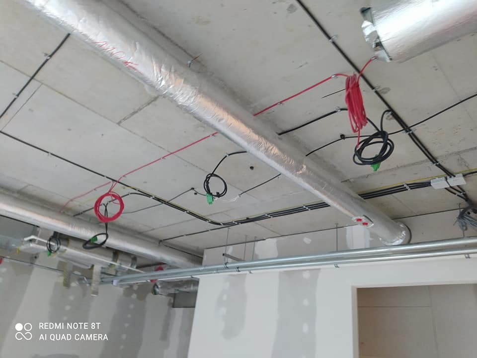 Instalacja wentylacyjna w budynku biurowym w trakcie montażu, widoczne rury owinięte folią aluminiową, kable elektryczne i sufit z betonu architektonicznego.