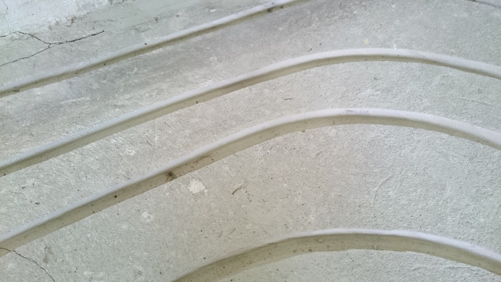 Świeżo wylana, szara betonowa podłoga z zatopionymi, zakrzywionymi rurkami instalacji grzewczej, widoczne pęknięcia i drobne niedoskonałości betonu.