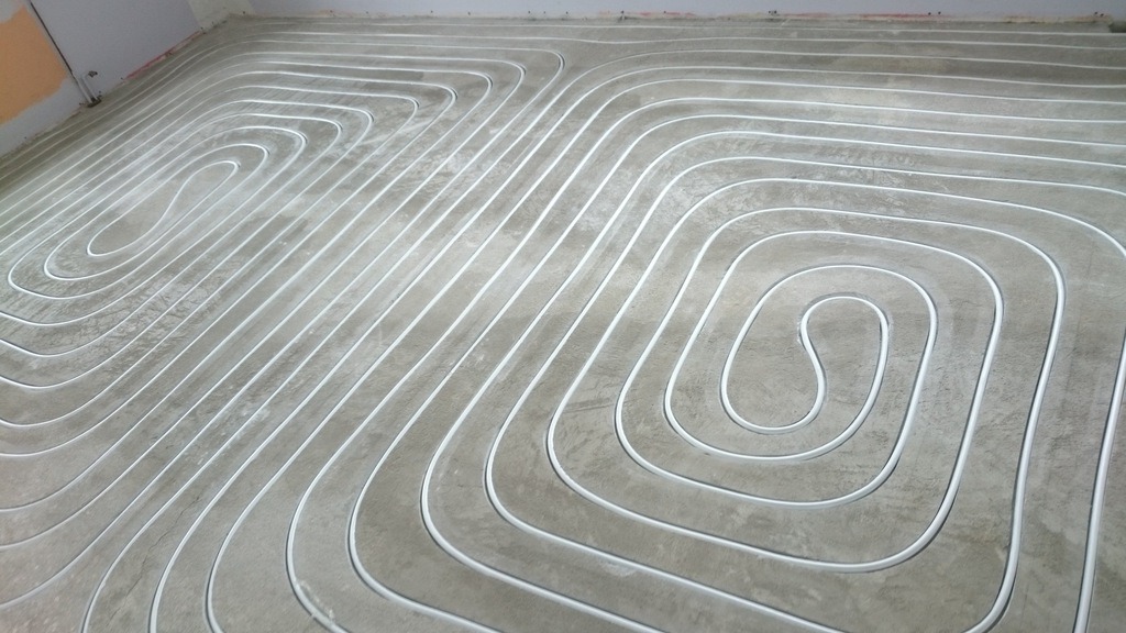 Białe rurki instalacji w systemie ogrzewania podłogowego, ułożone w spiralny wzór na szarej, betonowej wylewce, przed zalaniem. Widoczny fragment ściany z surowym tynkiem.