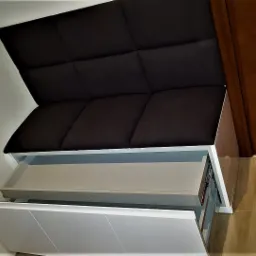 Pufa tapicerowana z szufladami w kolorze białym, z ciemnym siedziskiem i oparciem, ustawiona przy drewnianej ścianie.