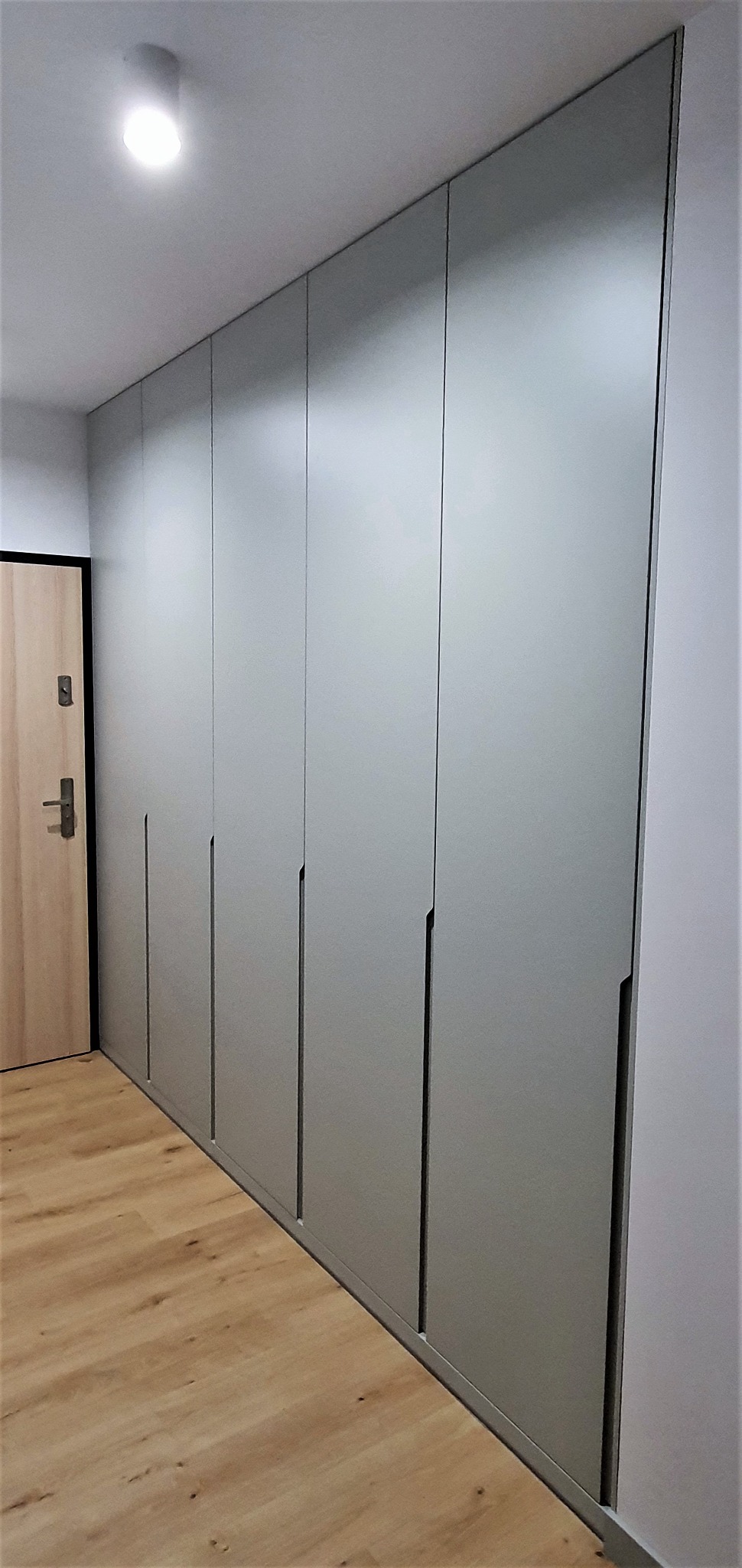 Długa, zabudowana szafa wnękowa z gładkimi, szarymi frontami i minimalistycznymi uchwytami, dopasowana do wysokości pomieszczenia, z widoczną podłogą z paneli oraz drzwiami wejściowymi po lewej...