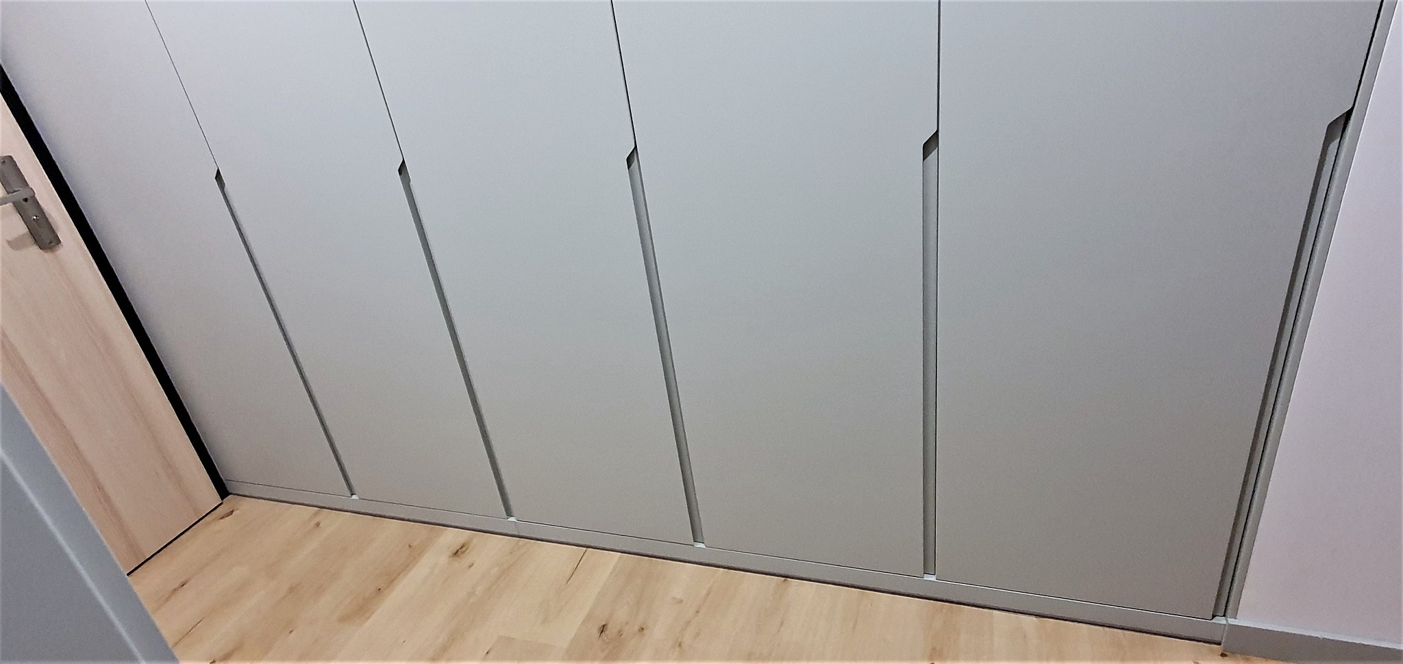 Szara szafa wnękowa z minimalistycznymi uchwytami w zabudowie, widoczna pod kątem na tle drewnianej podłogi.
