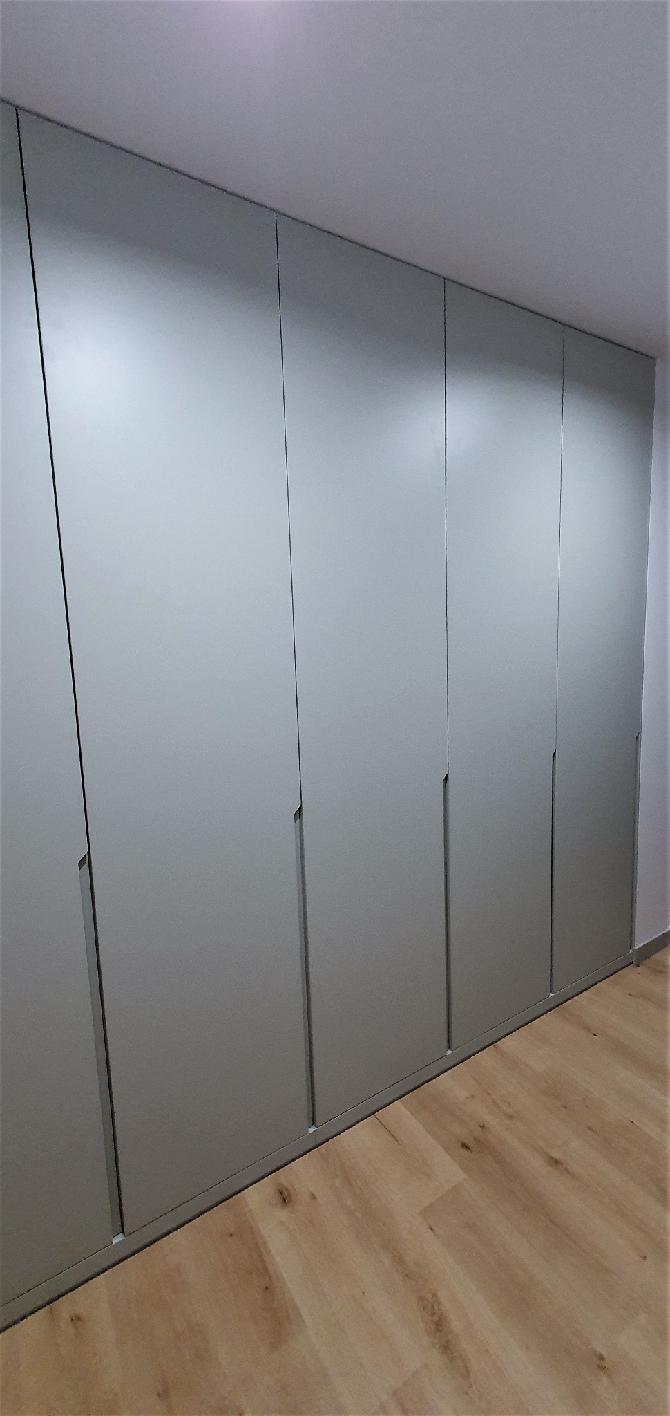 Długa, zabudowana szafa wnękowa z szarymi, gładkimi frontami i minimalistycznymi uchwytami, dopasowana do jasnej podłogi z paneli drewnopodobnych.