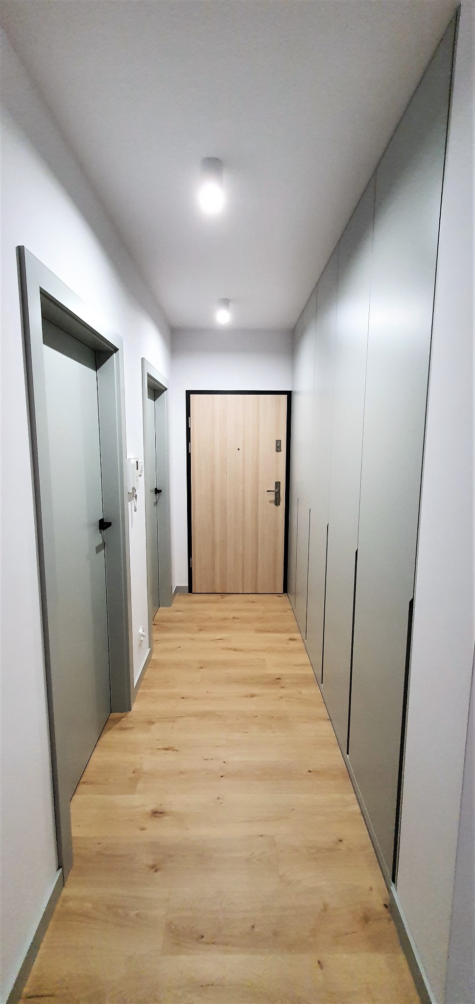 Długi korytarz z podłogą imitującą drewno, po prawej stronie rząd szaf w odcieniach szarości z minimalistycznymi uchwytami, po lewej dwie pary drzwi również w odcieniach szarości.