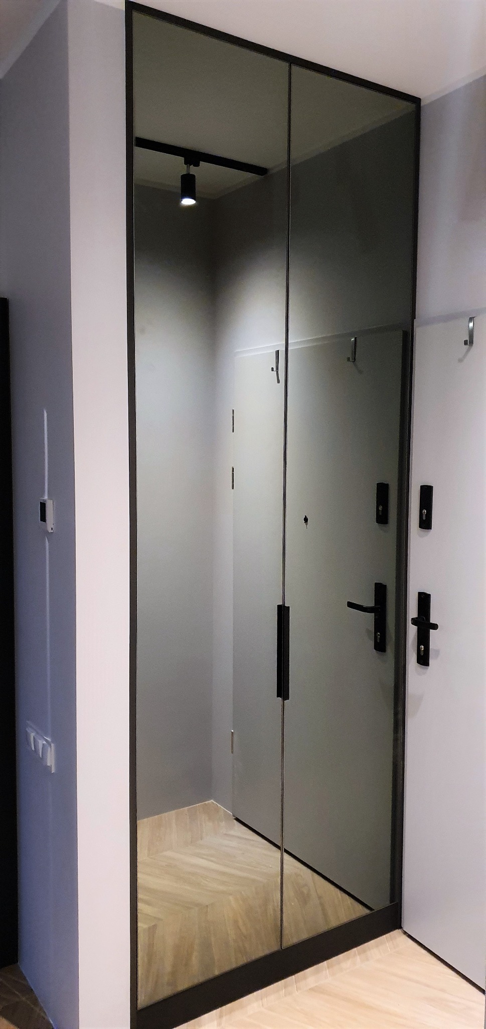 Dwu-drzwiowa szafa wnękowa z lustrzanymi frontami w czarnej ramie, odbijająca korytarz z parkietem ułożonym w jodełkę.