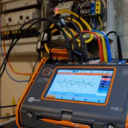 Miernik Sonel podłączony do rozdzielnicy elektrycznej, ekran pokazuje wykresy napięć i prądów, widoczne przewody pomiarowe w kolorach czarnym, czerwonym, żółtym i niebieskim.