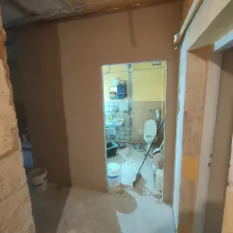 Ściany w trakcie gipsowania, widok na łazienkę z widoczną toaletą i umywalką, podłoga pokryta resztkami materiałów budowlanych, widoczny kubeł z zaprawą i narzędzia.